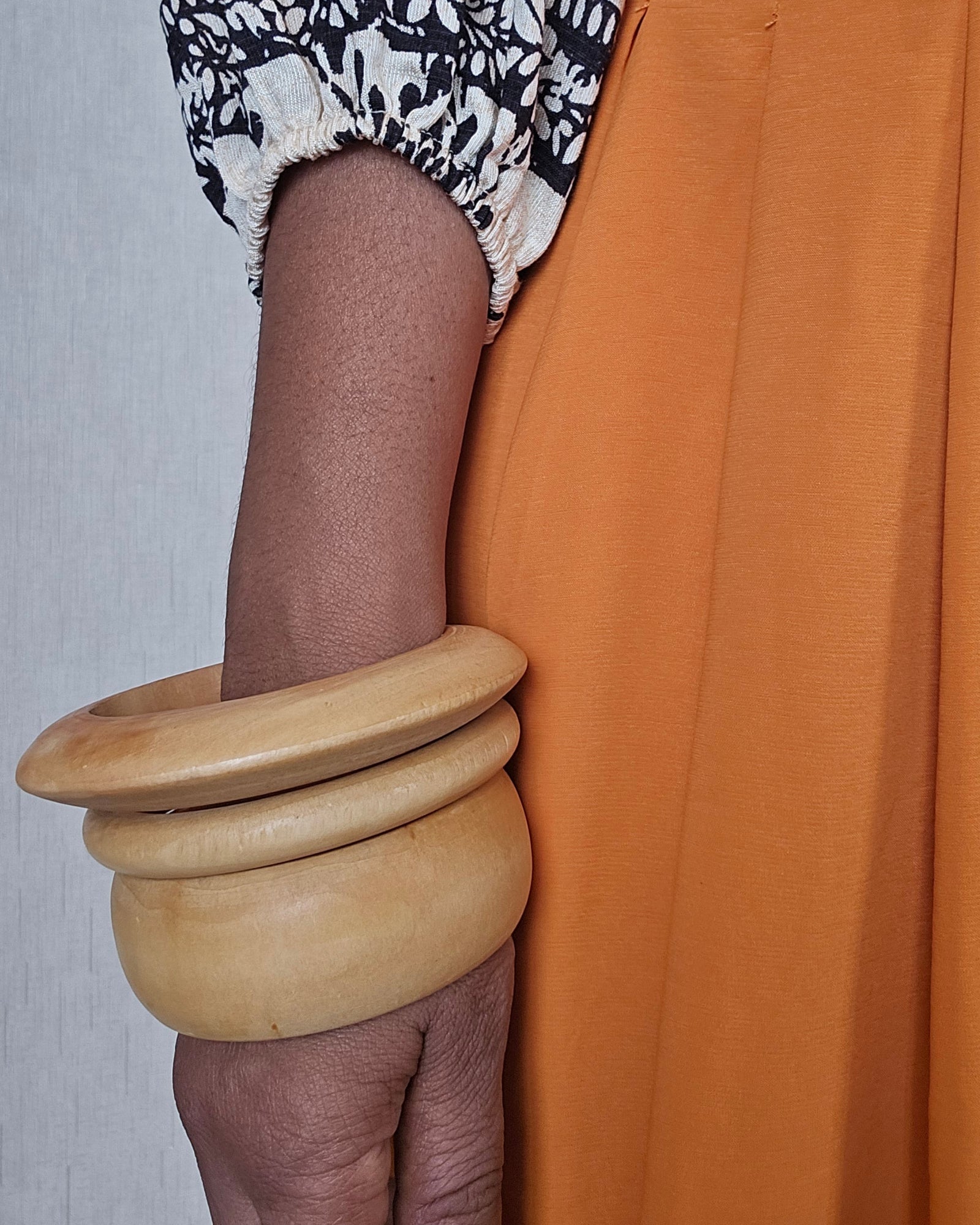 A close-up of a person wearing a wide, natural wood bangle on their wrist, paired with a black and white patterned blouse and an orange skirt.