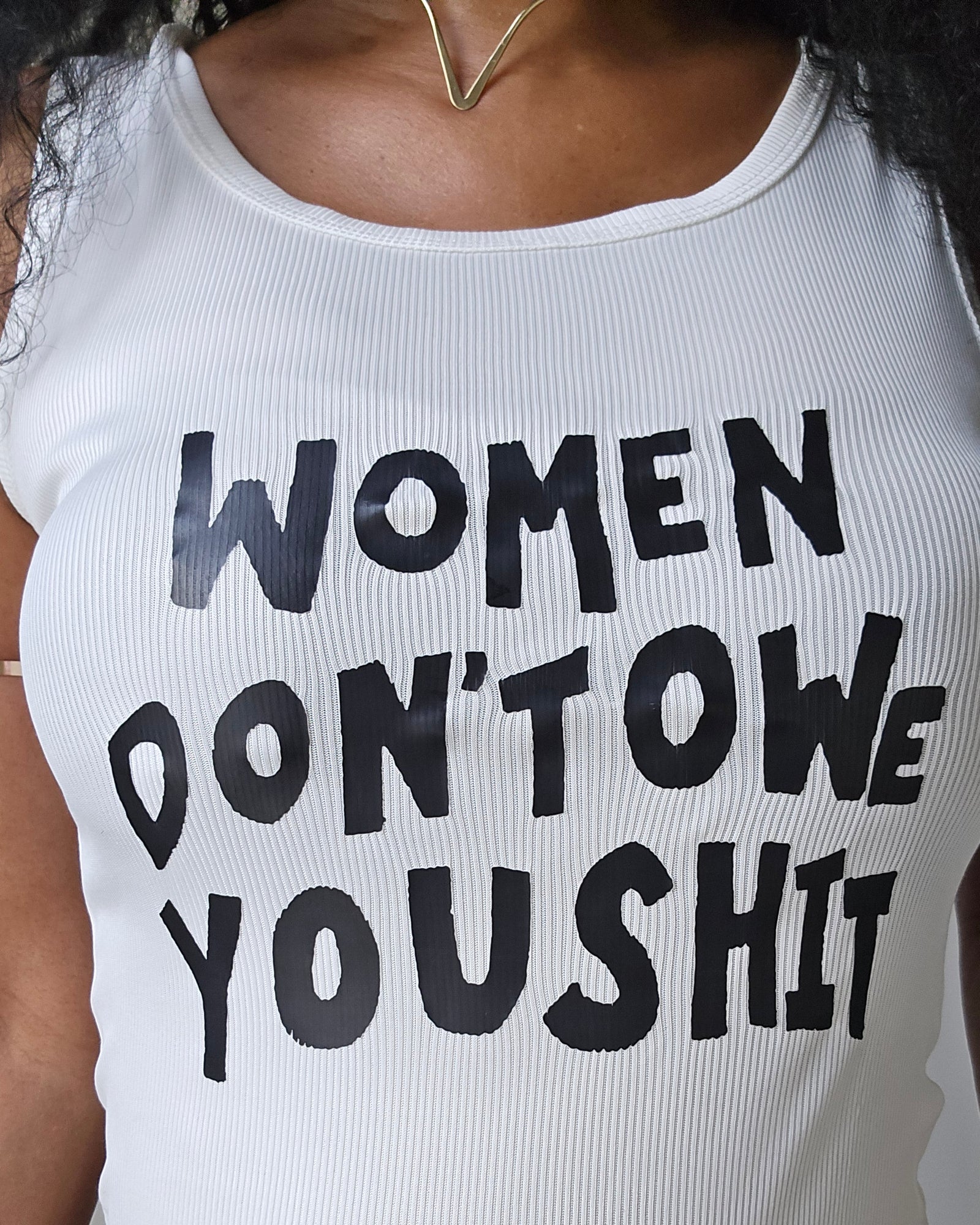 A close-up of a person wearing a white ribbed tank top with bold black text that reads 'WOMEN DON'TOWE YOUSHIT', paired with rust-colored shorts.