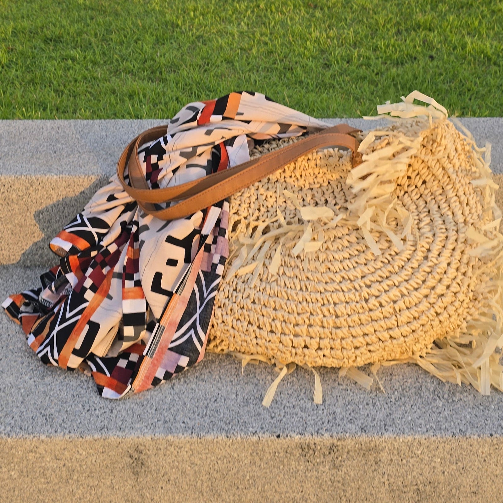 A straw woven tote bag with fringe and a multicolor patterned scarf with brown handles, resting on a concrete ledge in an outdoor setting with a grassy lawn and city view in the background.