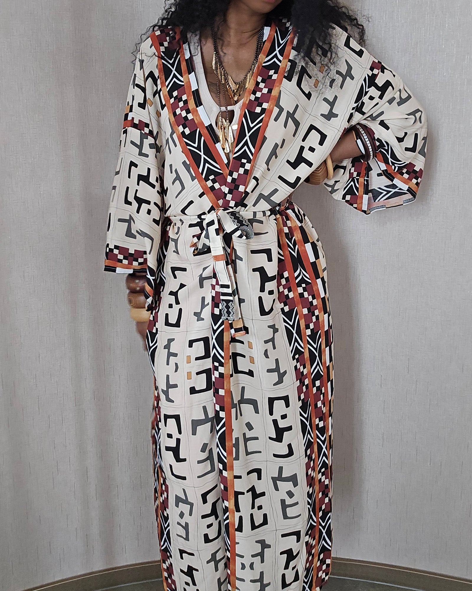 A woman wearing a long white robe with a bold black, red, and orange geometric pattern, paired with an orange headband, standing against a light gray textured wall.