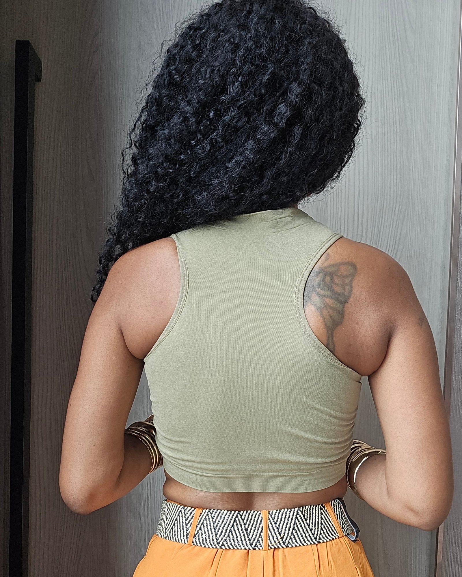 A woman viewed from behind wearing a light sage green ribbed tank top and bright orange wide-leg pants with a patterned waistband, standing indoors against a gray door.