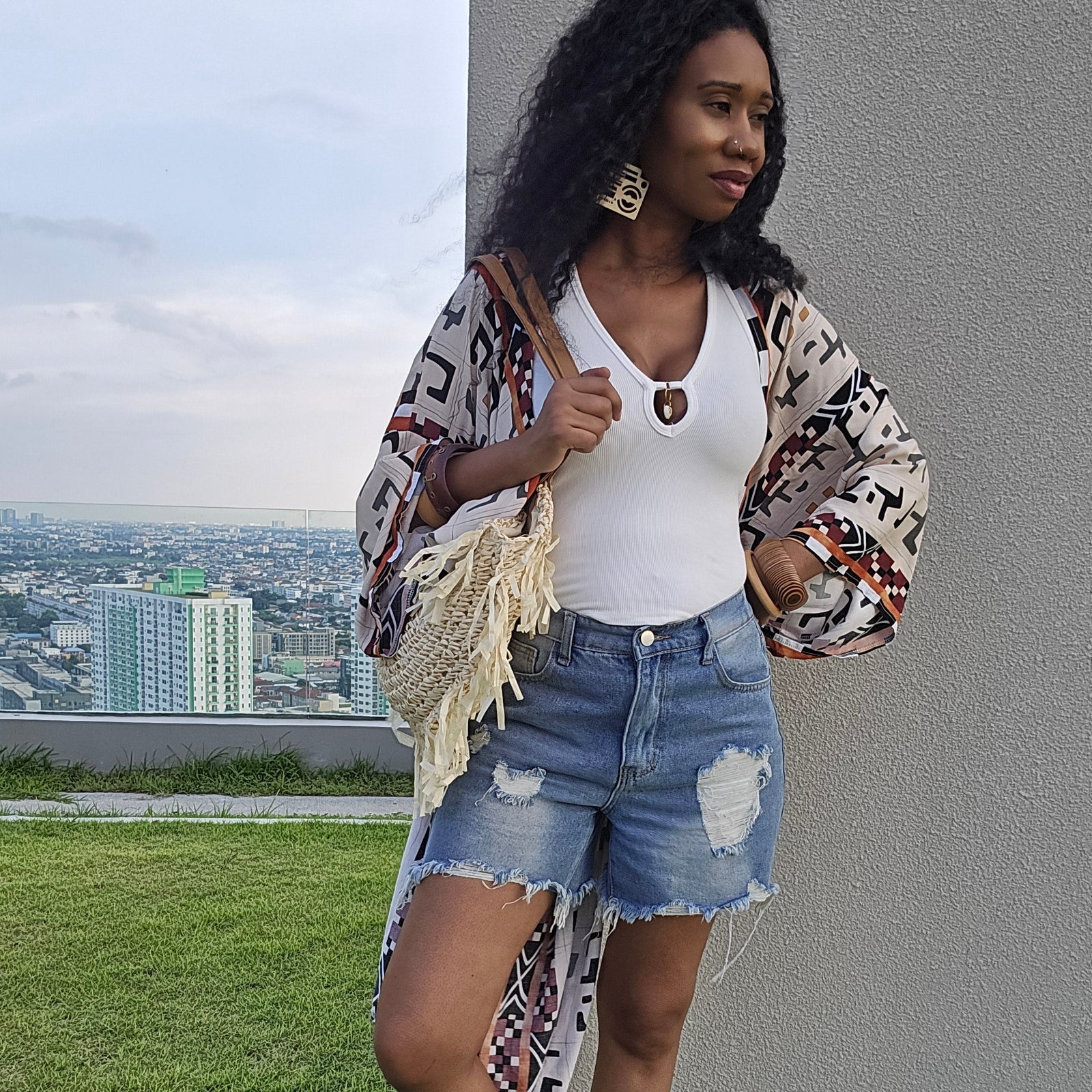 A woman stands on a rooftop lawn wearing a white tank top, distressed denim shorts, a long bohemian patterned cardigan, and flat sandals, with a city skyline and gray building wall in the background.