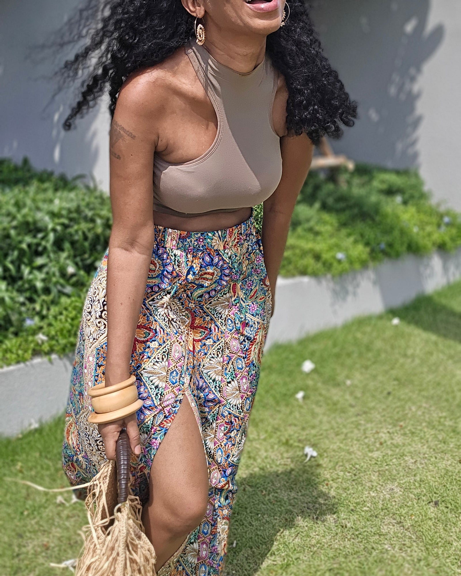 A woman laughing outdoors in a beige crop top and a colorful, patterned high-slit maxi skirt, holding a straw bag against a background of green grass and a gray wall.