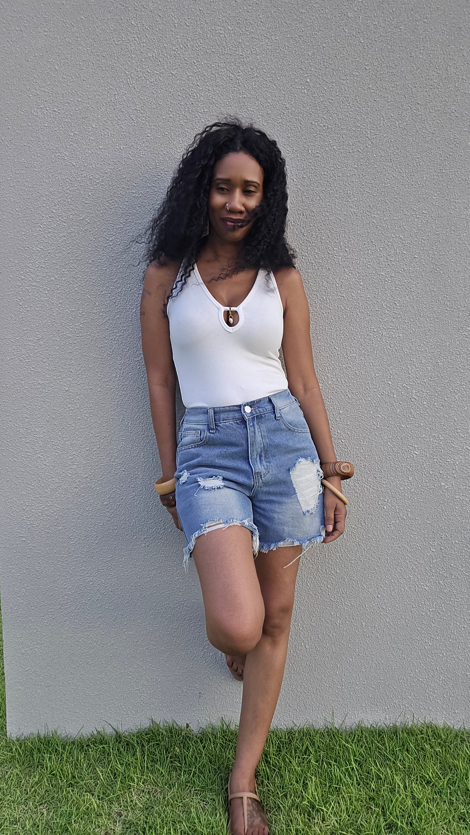 A woman posing outdoors against a gray wall and green grass, wearing a white sleeveless top and distressed denim shorts.
