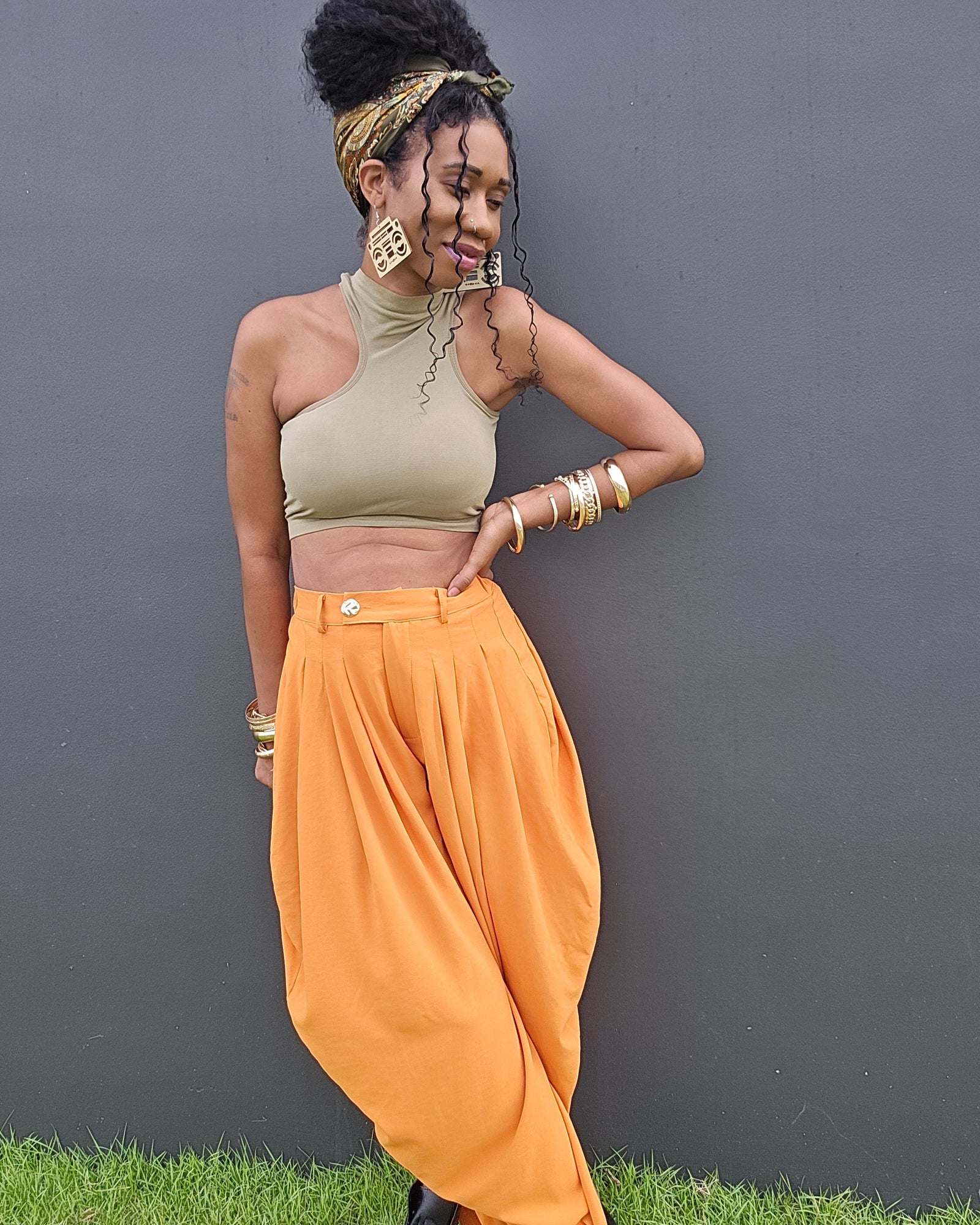 A woman posing outdoors against a dark gray wall on a patch of green grass, wearing a beige halter crop top, vibrant orange harem-style pants, and black boots.