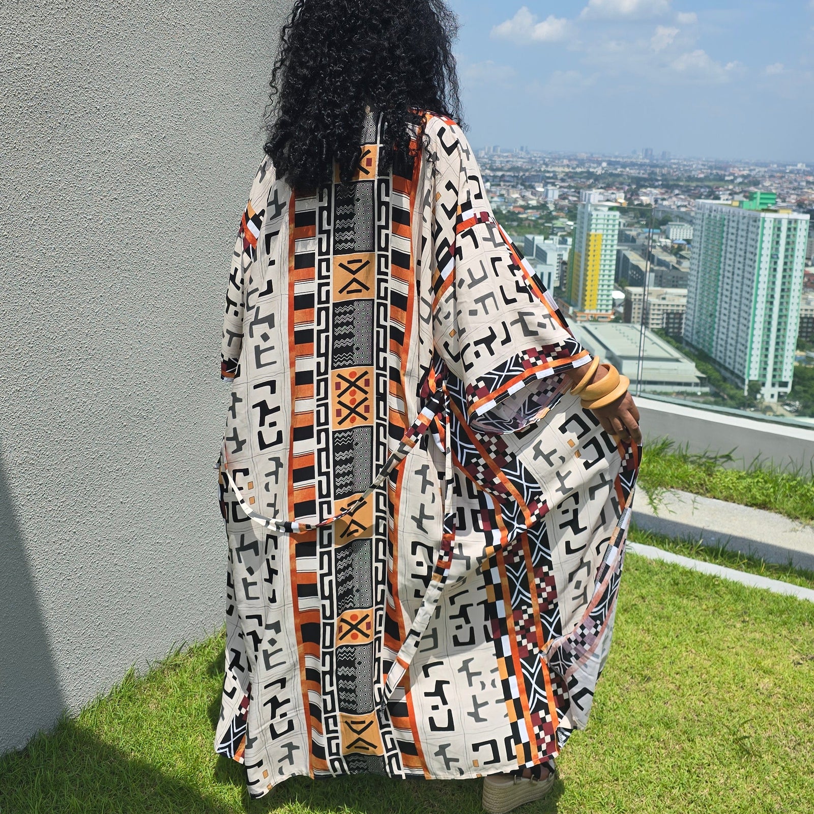 A person viewed from behind wearing a long, flowing kaftan-style robe with a bold geometric tribal pattern in black, orange, white, and gray. The person is standing on a grassy rooftop terrace next to a gray concrete wall, looking out over a city skyline under a blue sky with white clouds.