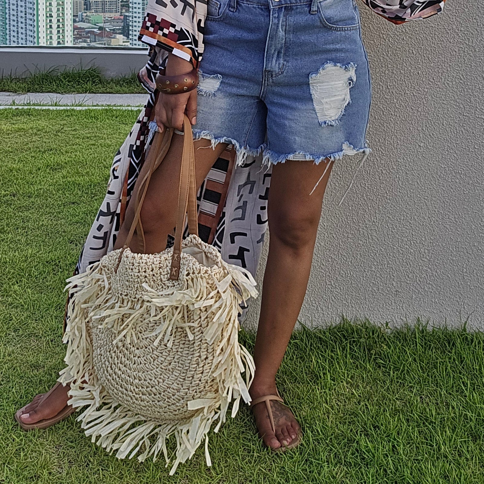 A woman stands outdoors on a grassy rooftop wearing a white tank top, distressed denim shorts, a long patterned open-front cardigan, and carrying a large woven straw bag.