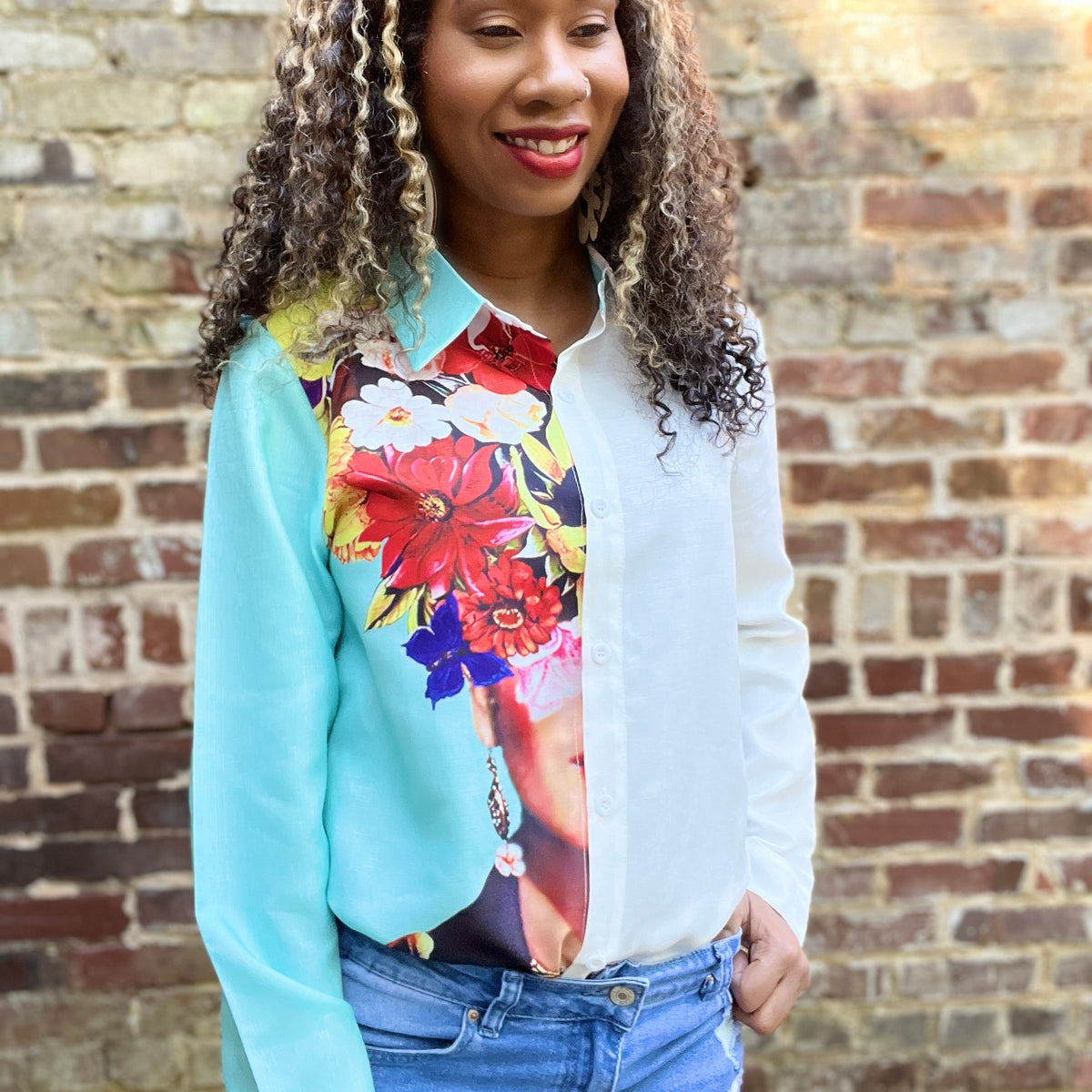 A woman wearing a long-sleeve button-up blouse with a white collar and front panel, layered over a turquoise cardigan featuring a large multicolor floral print. She is standing in front of a brick wall.