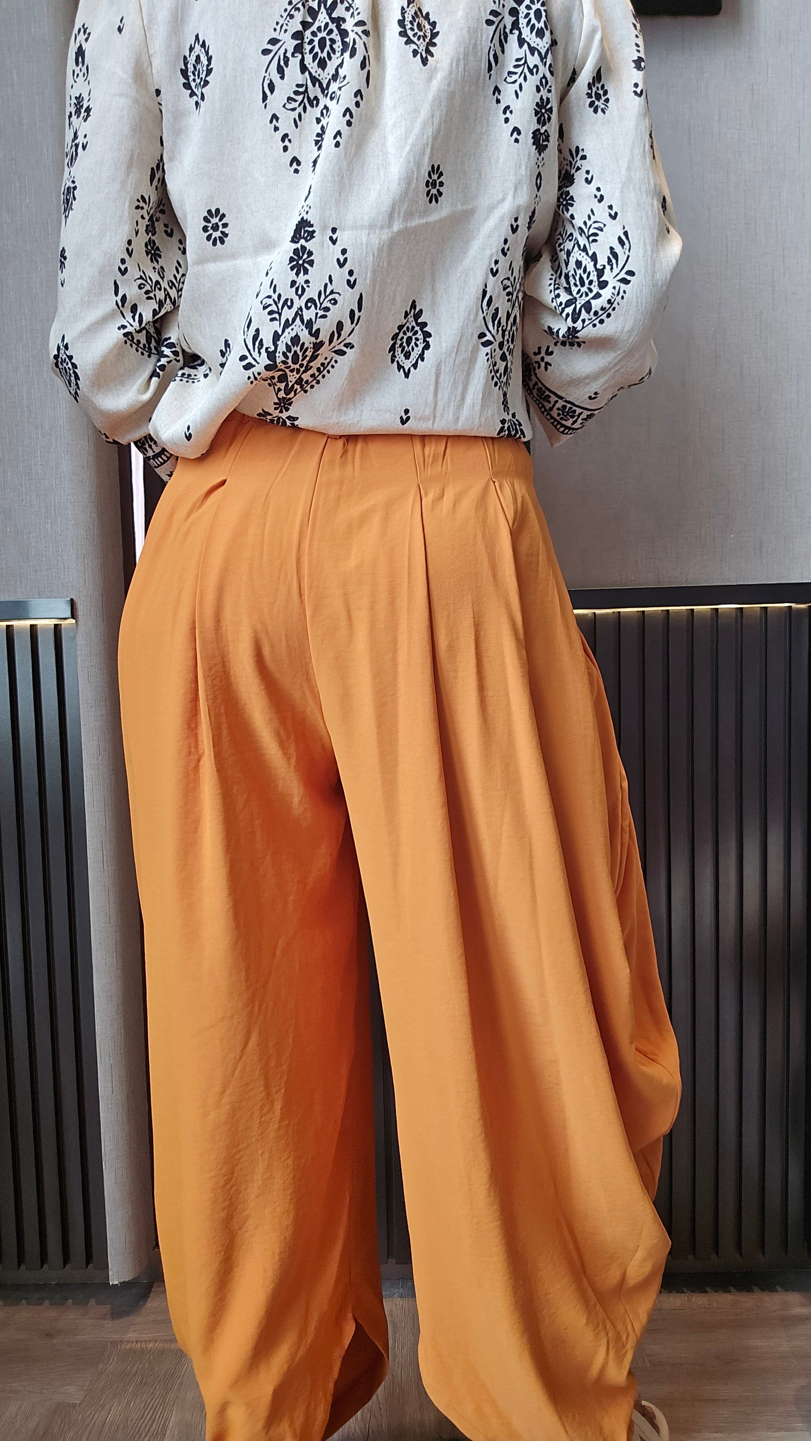 A person shown from the back wearing loose, wide-leg orange pants and a white long-sleeve blouse with black floral print, standing indoors in front of a dark slatted wall.