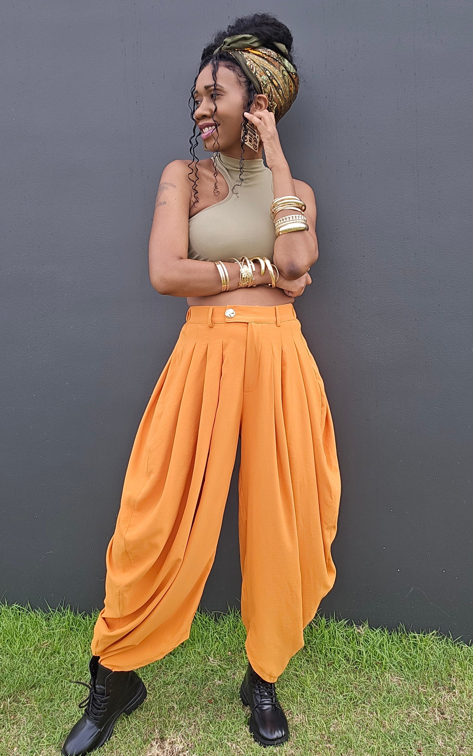 A woman posing outdoors in front of a dark gray wall on a patch of green grass, wearing a beige one-shoulder top, bright orange harem-style pants, a gold and black patterned headwrap, and black lace-up boots.
