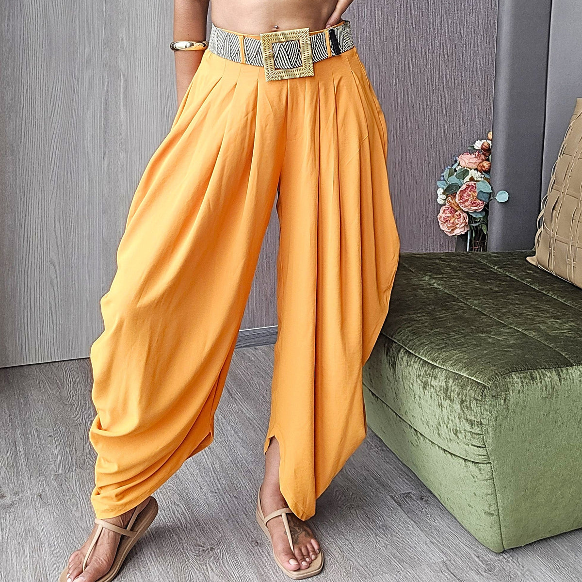 A person wearing loose, draped harem-style pants in a warm peachy orange color, paired with a patterned belt and sandals, standing indoors against a gray wall.