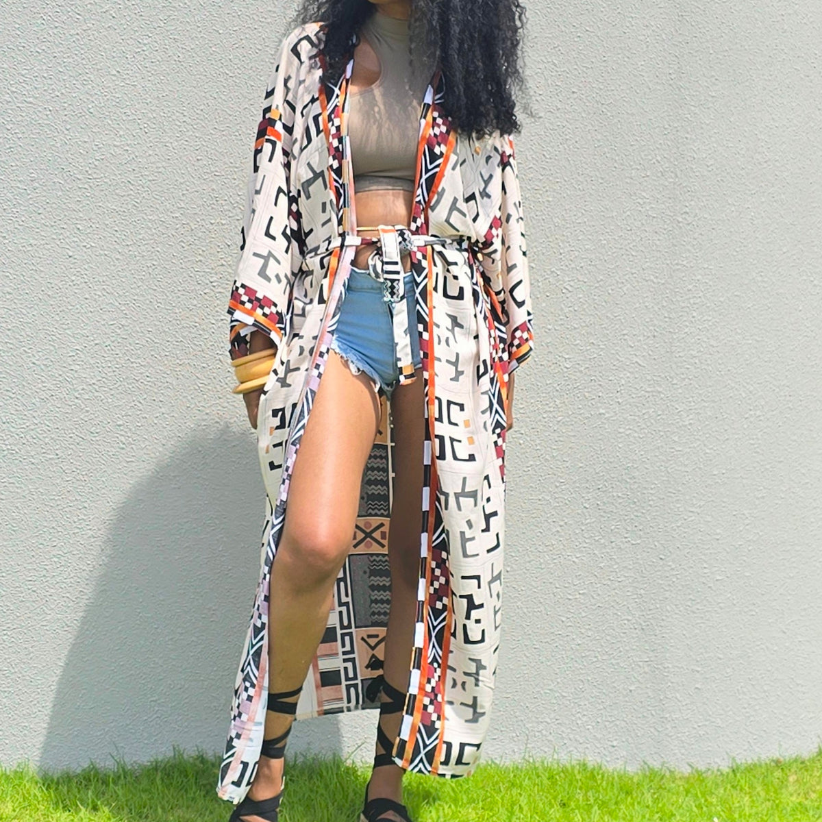 A woman with long curly black hair stands outdoors on grass in front of a large light gray concrete structure, wearing a long white open-front coat with a bold multicolor geometric pattern, a beige crop top, denim shorts, and black sandals.