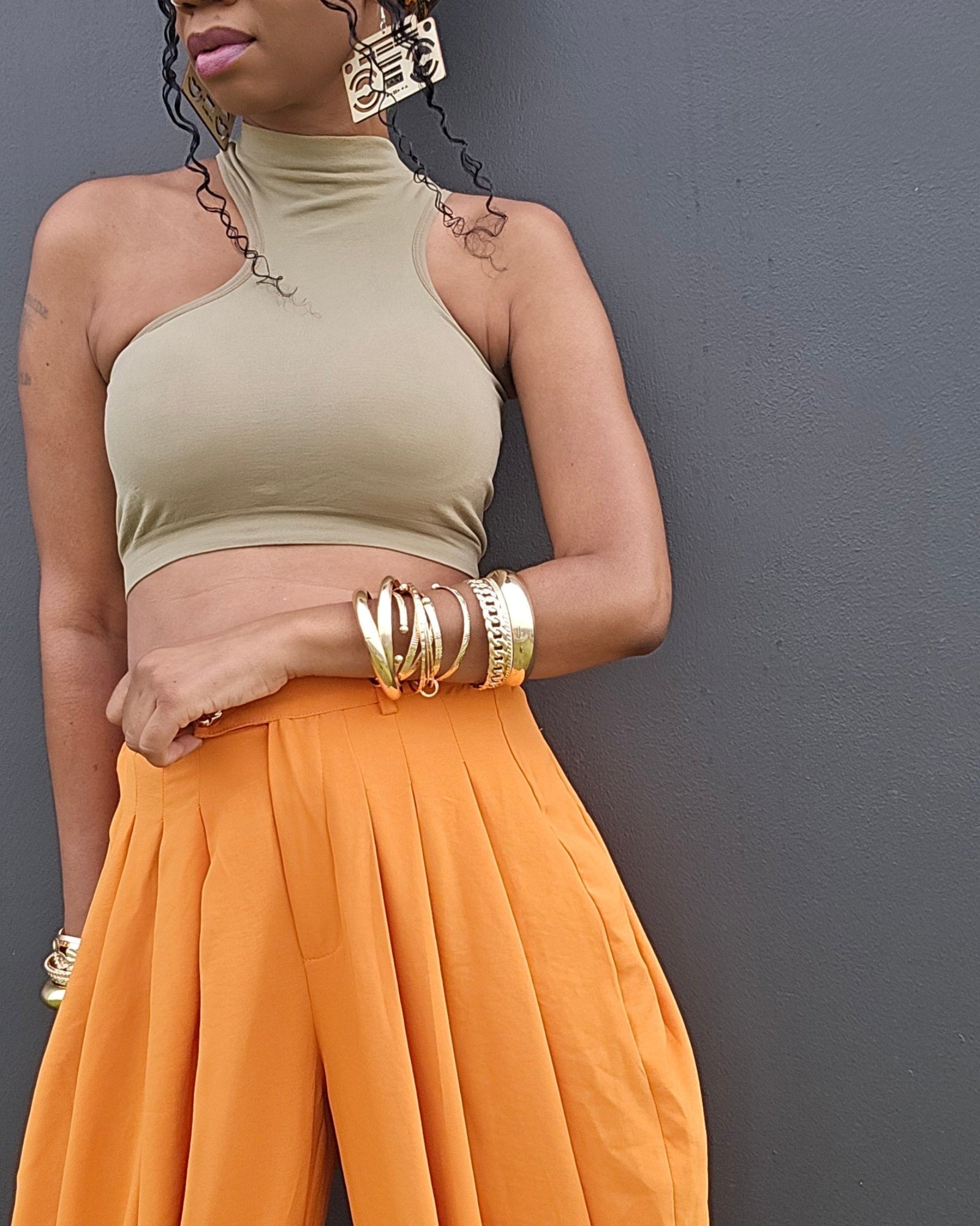 A woman wearing a beige halter crop top and bright orange wide-leg pants, standing against a dark gray wall.
