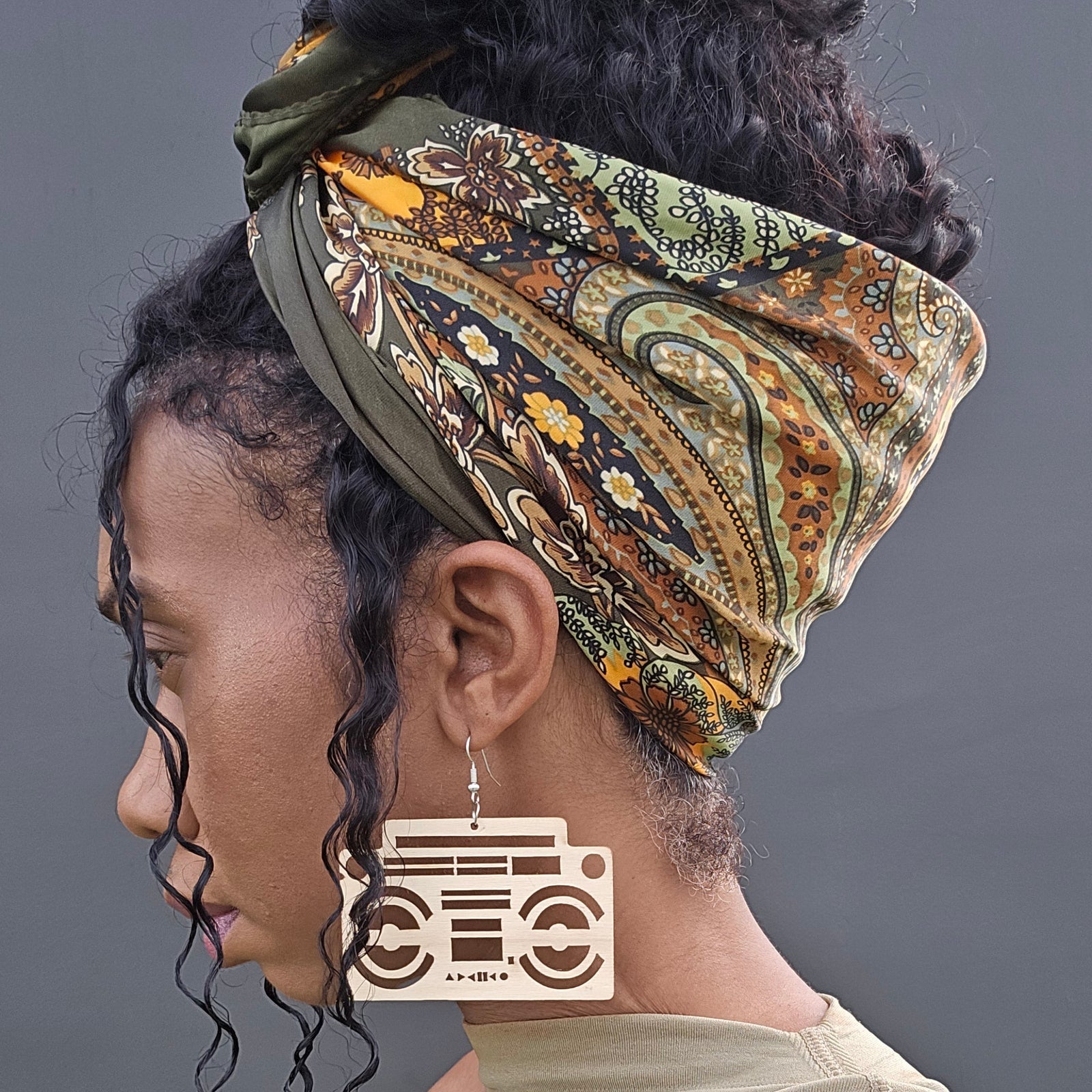 A woman shown in profile wearing a patterned headwrap in olive green, brown, and gold tones, along with large rectangular wooden earrings shaped like retro boomboxes, against a dark gray studio background.