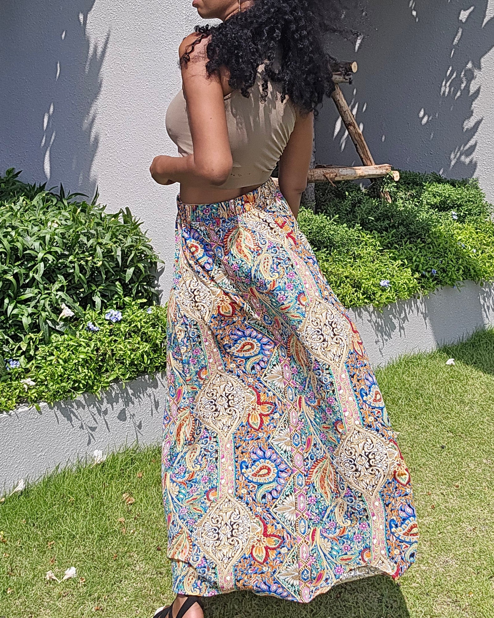 A woman wearing a beige sleeveless top and a long, colorful bohemian-patterned maxi skirt stands outdoors on a grassy lawn in front of a gray wall with green plants.