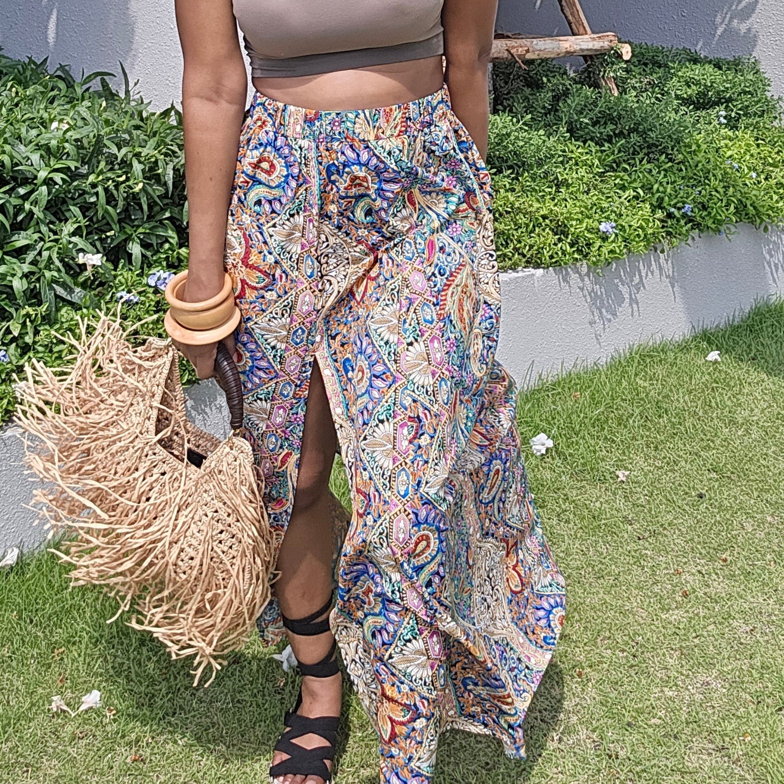 A woman stands outdoors on a grassy lawn wearing a taupe halter crop top and a colorful, patterned asymmetrical wrap skirt. She is holding a straw hat and wearing black strappy sandals.