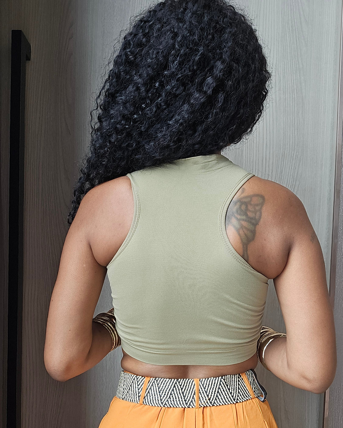 A woman viewed from behind wearing a light sage green ribbed tank top and bright orange wide-leg pants with a patterned waistband, standing indoors against a gray door.