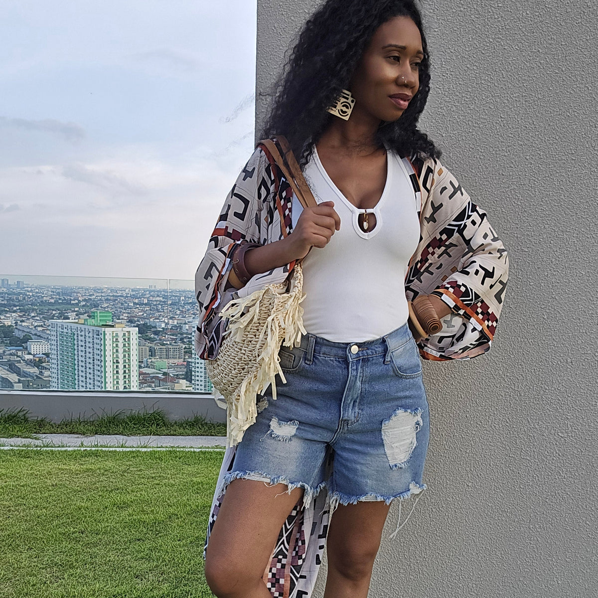 A woman stands on a rooftop lawn wearing a white tank top, distressed denim shorts, a long bohemian patterned cardigan, and flat sandals, with a city skyline and gray building wall in the background.