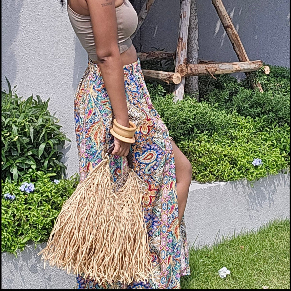 A woman stands on a grassy lawn beside a gray wall and small tree, wearing a beige crop top and a long multicolor bohemian skirt, holding a straw bag.