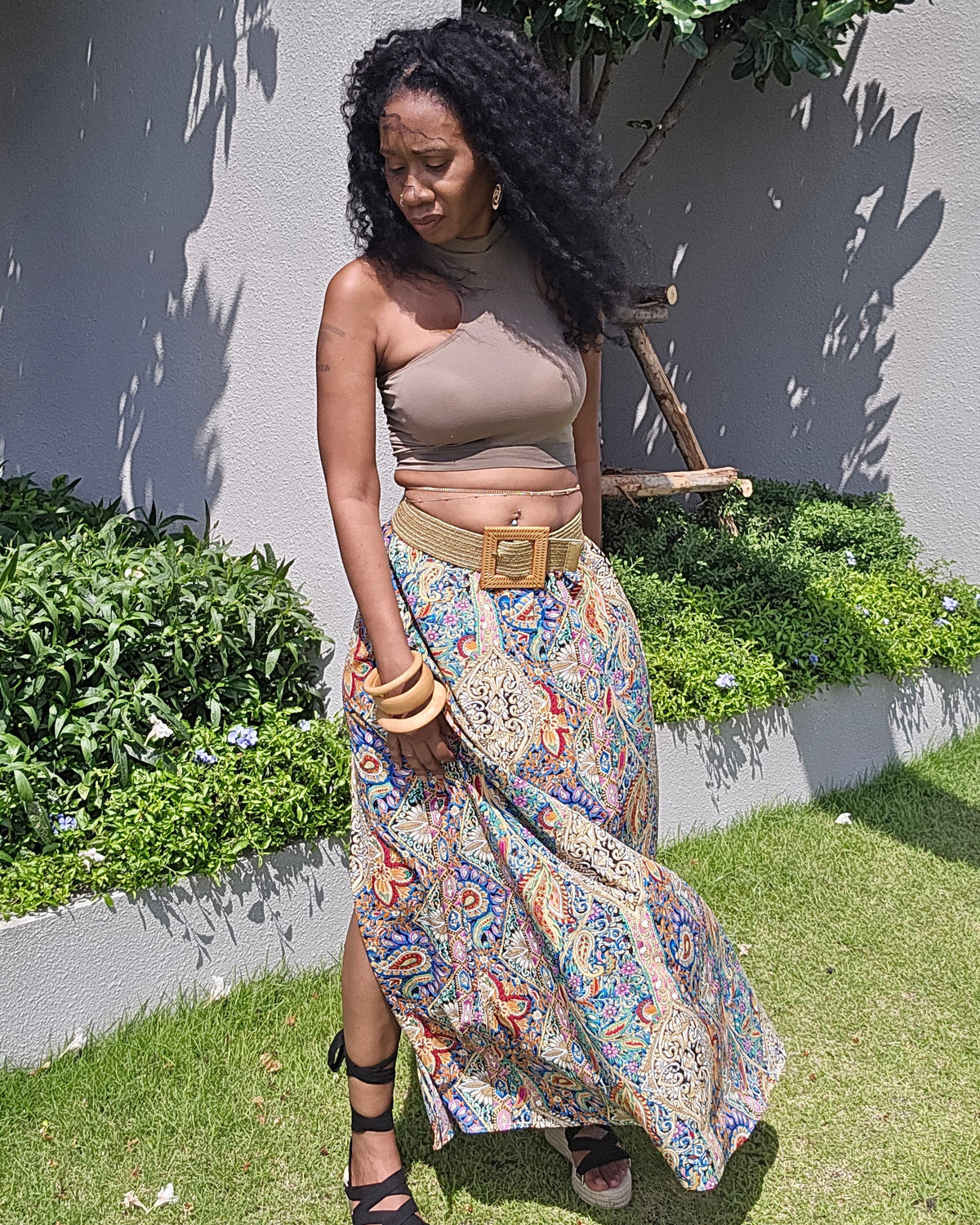 A woman stands outdoors on a grassy lawn in front of a gray wall and green plants, wearing a beige strapless crop top, a long multicolor paisley skirt with a gold belt, and black strappy sandals.