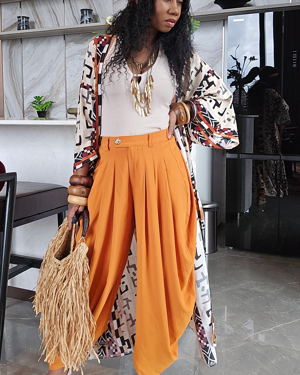 A woman models a bohemian-style outfit featuring bright orange pleated wide-leg pants, a white top, a patterned open-front cardigan, an orange headband, and a straw bag, set against a modern interior background.