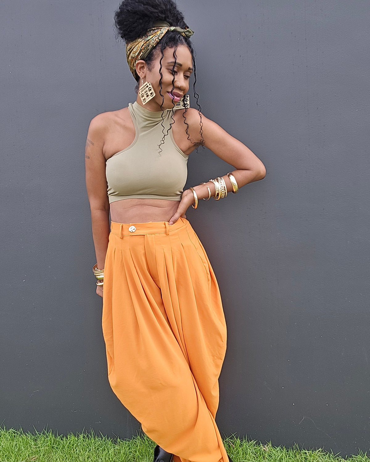 A woman posing outdoors against a dark gray wall on a patch of green grass, wearing a beige halter crop top, vibrant orange harem-style pants, and black boots.