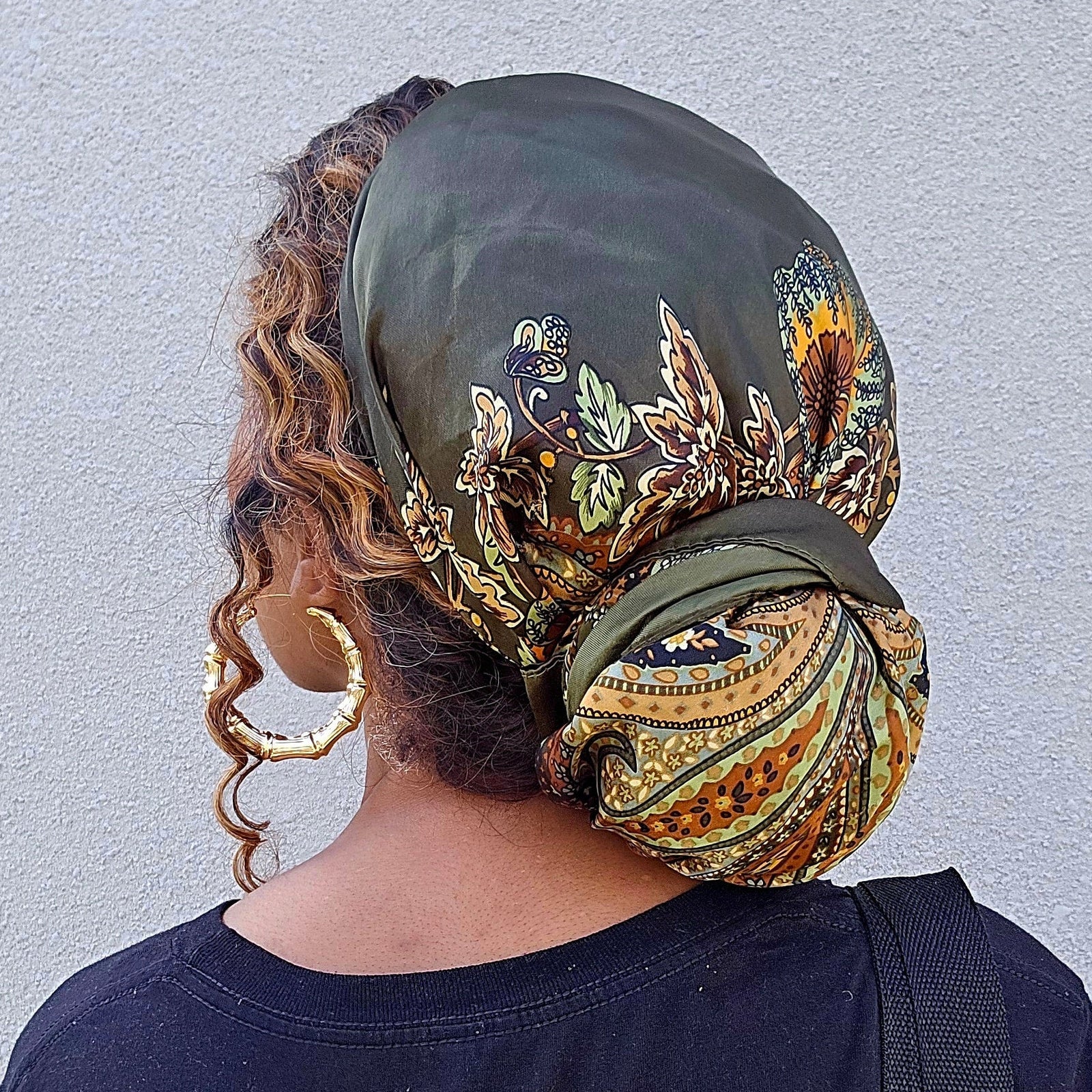 A woman shown from behind wearing a dark olive green headscarf with a colorful paisley pattern, styled in a bun. She is wearing a dark navy t-shirt and a yellow skirt, and carrying a green patterned shoulder bag against a light gray textured wall.