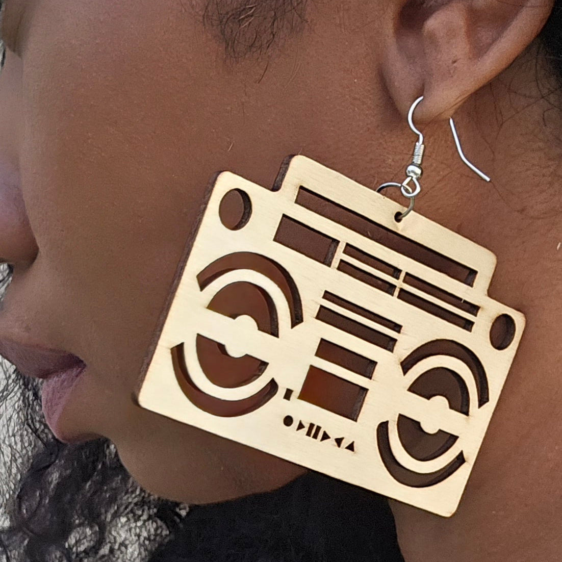 A close-up of a person wearing large laser-cut wooden boombox earrings with silver fishhook wires.